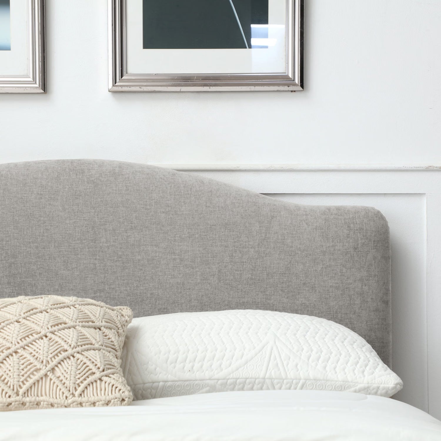 A close-up of a gray upholstered headboard in a bedroom setting, with a white pillow, a cream textured accent pillow, and framed wall art above.