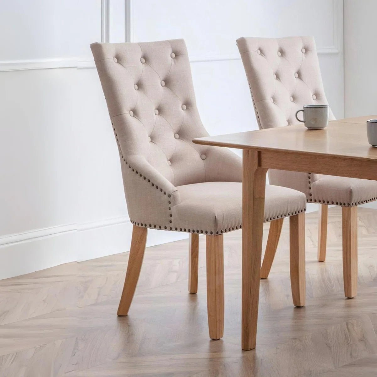 Two upholstered dining chairs with tufted backs and nailhead trim, shown beside a light wood dining table in a bright room with wood flooring.