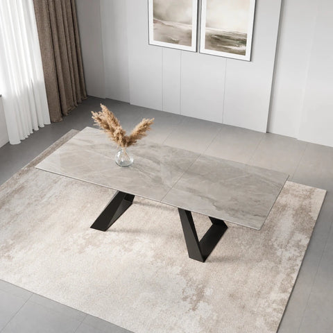 A modern rectangular Vortex dining table with a light gray stone-look Ceramic top and Dark Grey angular metal legs, styled on a beige rug in a minimalist room with a vase of dried grass.
