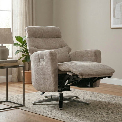 A beige upholstered recliner chair with an extended footrest, shown in a styled living room setting with a side table, lamp, plant, and patterned rug.