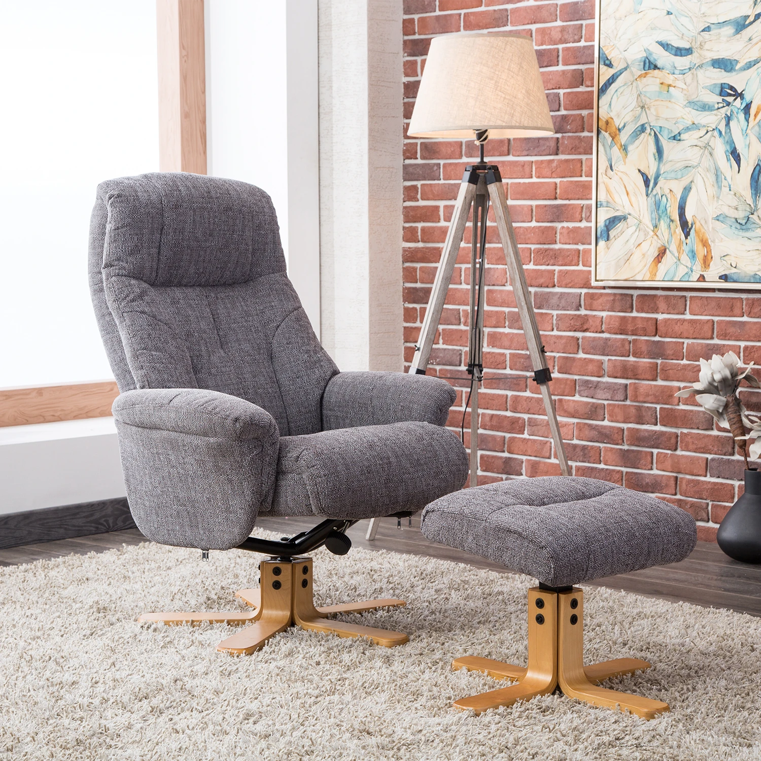 Gray fabric Muscat recliner chair with matching ottoman in a styled room setting featuring a brick wall, tripod floor lamp, and abstract wall art.