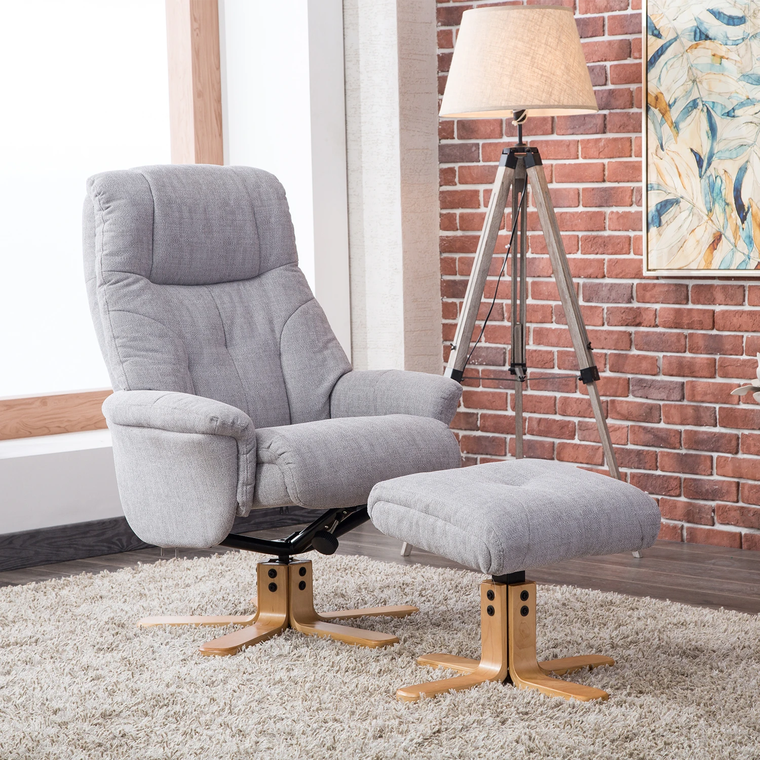 A light gray upholstered muscat recliner chair with matching ottoman in a styled room setting featuring a brick wall, tripod floor lamp, and shag rug.