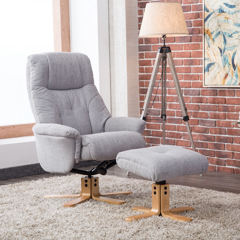 A light gray upholstered muscat recliner chair with matching ottoman in a styled room setting featuring a brick wall, tripod floor lamp, and shag rug.