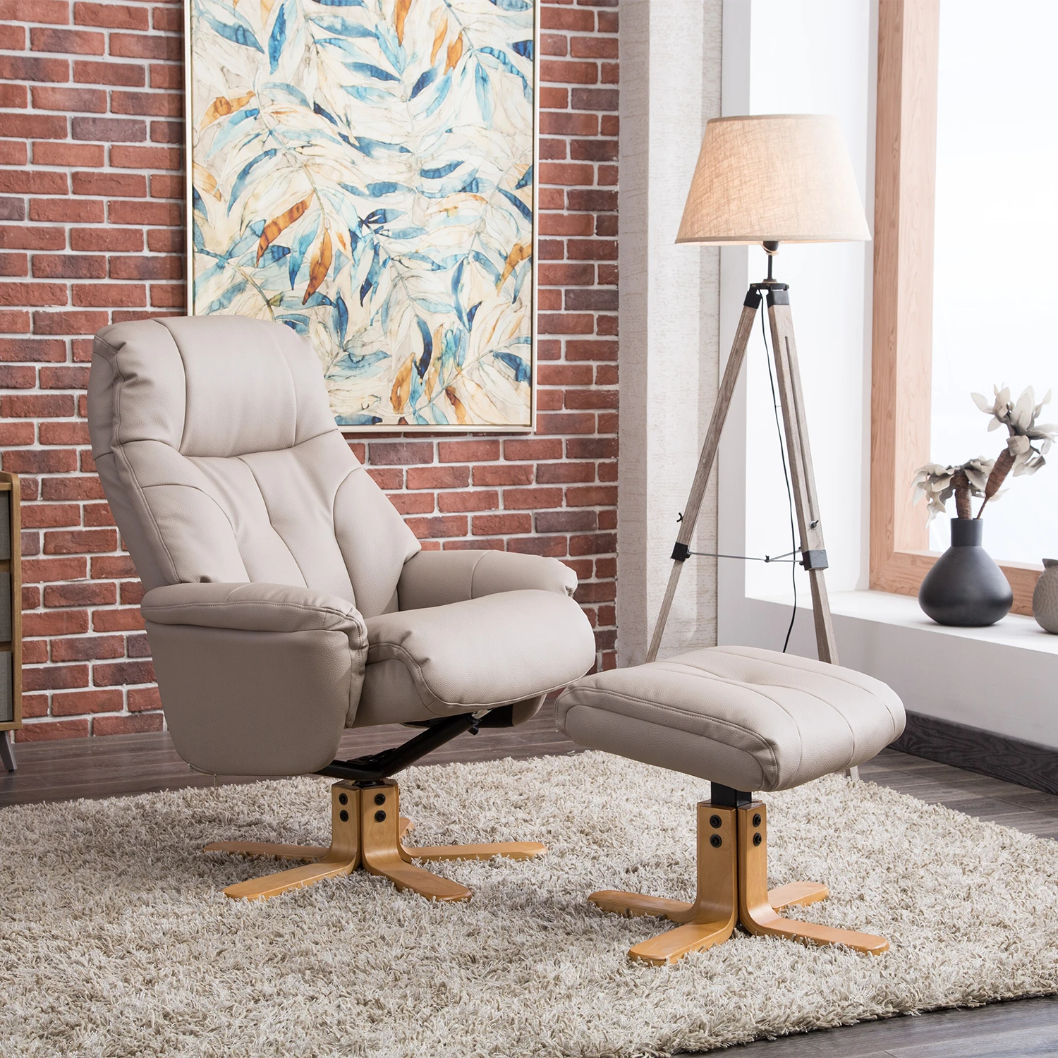 A beige upholstered recliner chair with a matching ottoman is shown in a styled living room setting with a brick wall, abstract painting, floor lamp, rug, and window.