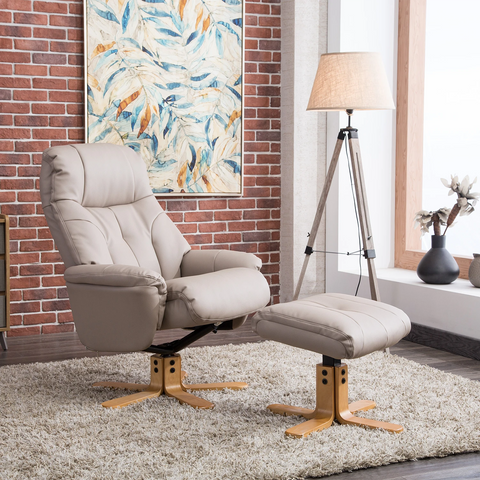 A beige upholstered recliner chair with a matching ottoman is shown in a styled living room setting with a brick wall, abstract painting, floor lamp, rug, and window.