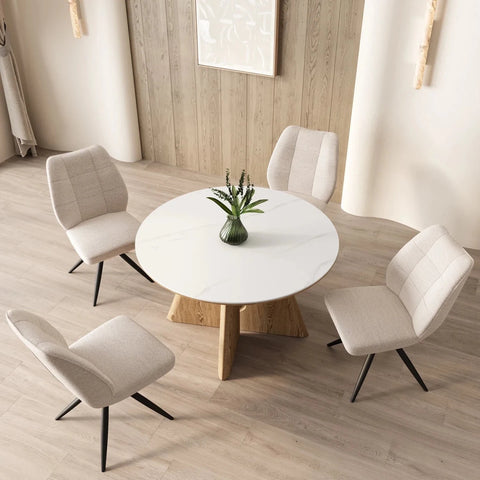 A modern Reese dining set featuring a round white marble effect ceramic top table with a natural wood pedestal base, surrounded by four upholstered dining chairs in a light beige fabric with black metal legs, set in a bright dining room.
