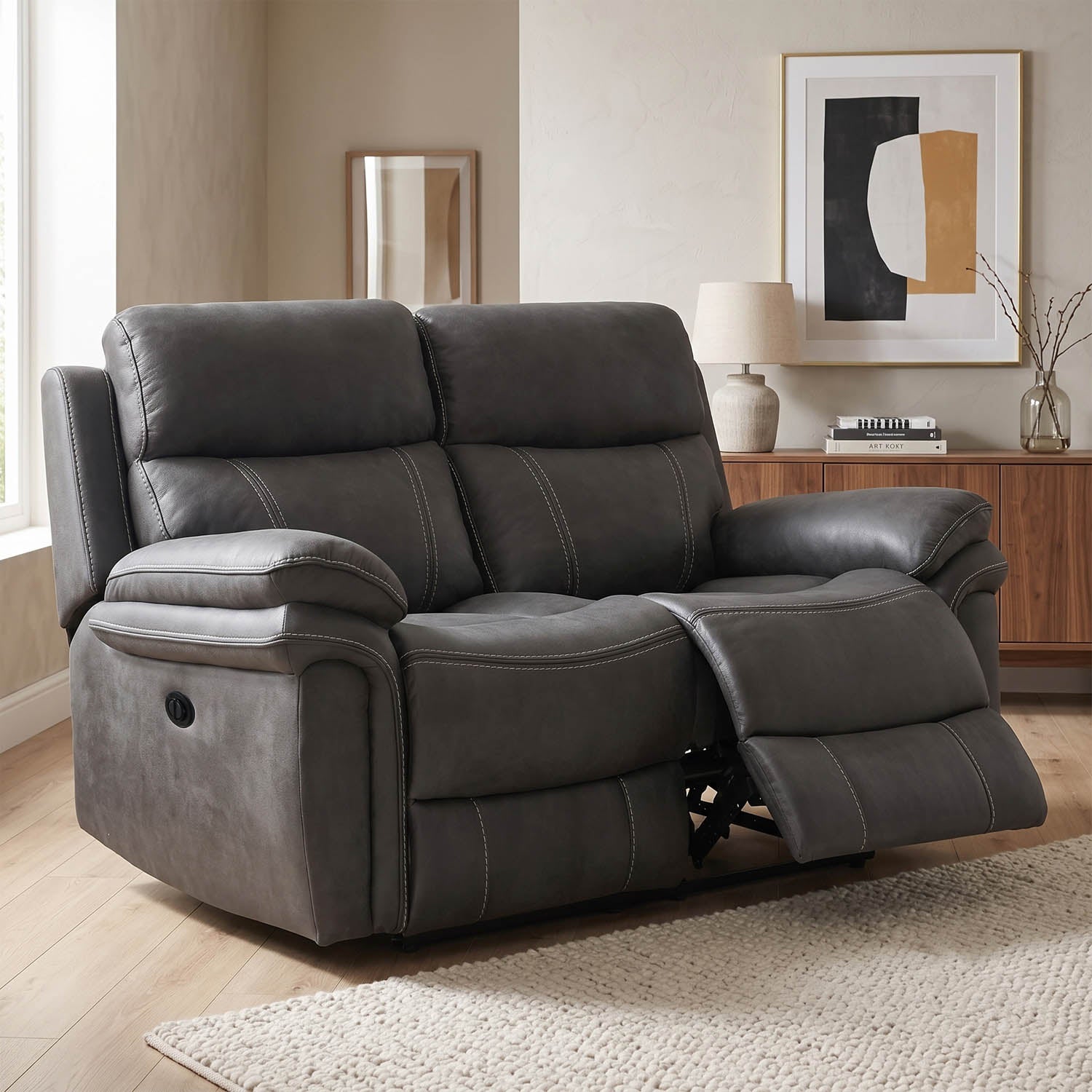 A dark gray upholstered reclining loveseat shown in a staged living room with wood flooring, a sideboard, lamp, and framed wall art.