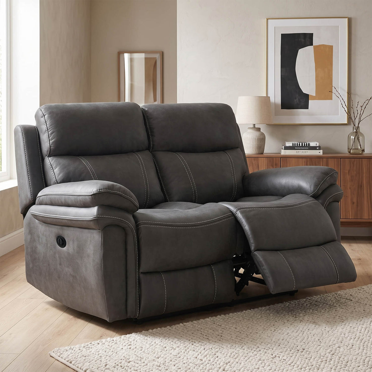 A dark gray upholstered reclining loveseat shown in a staged living room with wood flooring, a sideboard, lamp, and framed wall art.