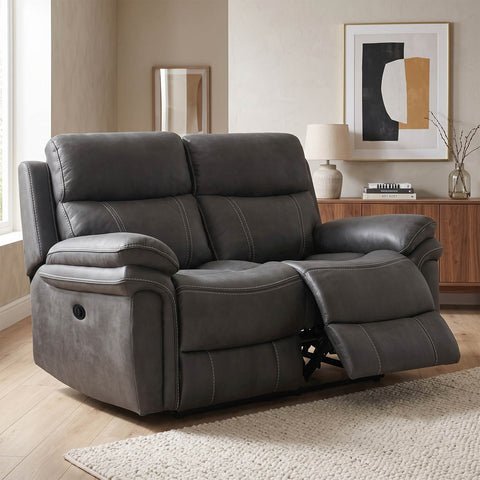 A dark gray upholstered reclining loveseat shown in a staged living room with wood flooring, a sideboard, lamp, and framed wall art.