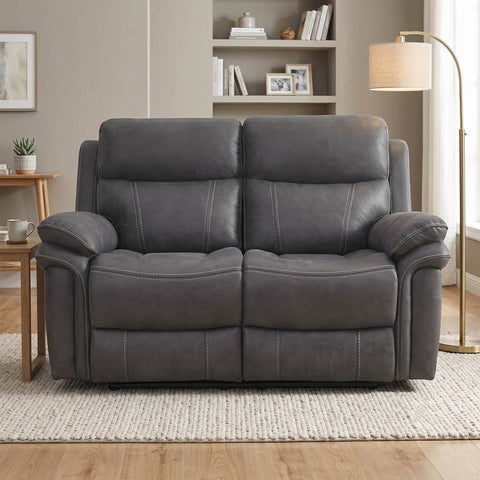 A dark gray upholstered reclining loveseat with padded armrests and channel tufting, shown in a staged living room setting with a side table, rug, floor lamp, and bookshelves in the background.