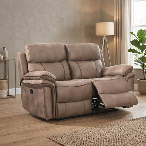 A taupe upholstered reclining loveseat shown in a staged living room with wood flooring, a floor lamp, side table, curtains, and a potted plant.