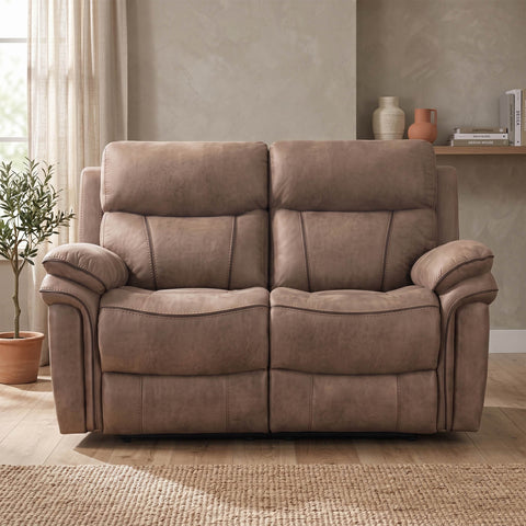 A two-seat reclining loveseat in a warm taupe-brown faux leather, shown in a styled living room setting with a potted plant, textured rug, and wall decor.