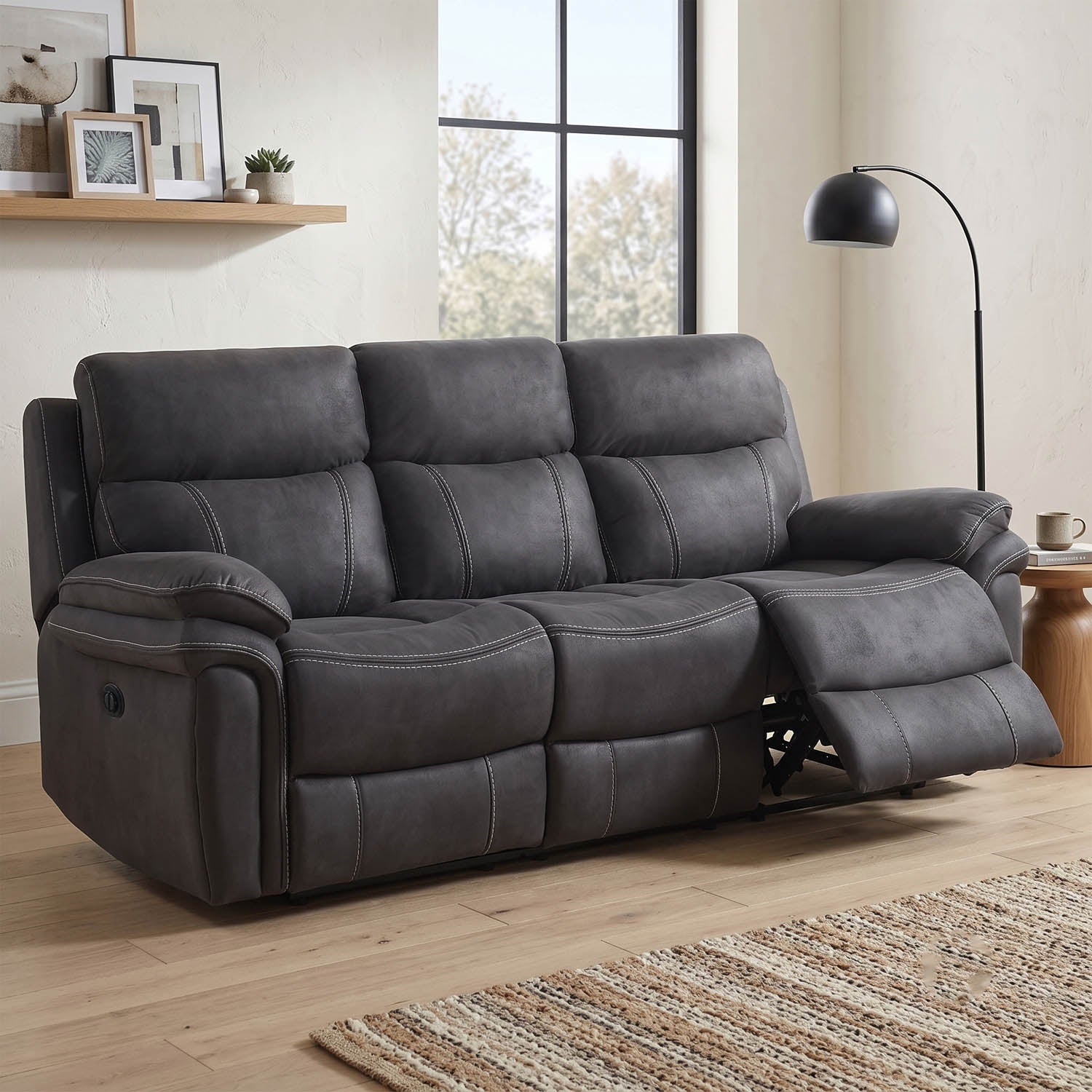 A dark gray three-seat reclining sofa with a pulled-out right side cushion, shown in a bright living room with wood flooring, a window, a floor lamp, and a small side table.