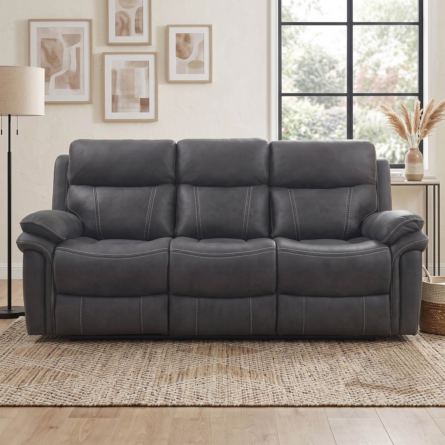 A dark gray three-seat reclining sofa with padded armrests and channel-tufted back cushions, shown in a staged living room setting with a woven rug, wall art, and a window.