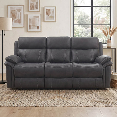 A dark gray three-seat reclining sofa with padded armrests and channel-tufted back cushions, shown in a staged living room setting with a woven rug, wall art, and a window.