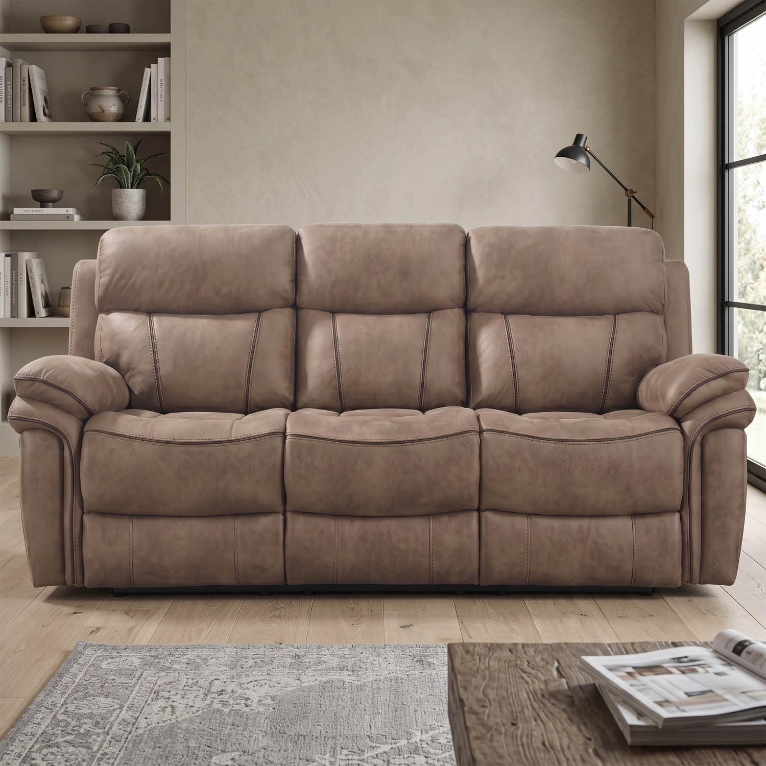 A three-seat reclining sofa upholstered in taupe-brown plush fabric, shown in a staged living room setting with a rug, bookshelf, lamp, and window.