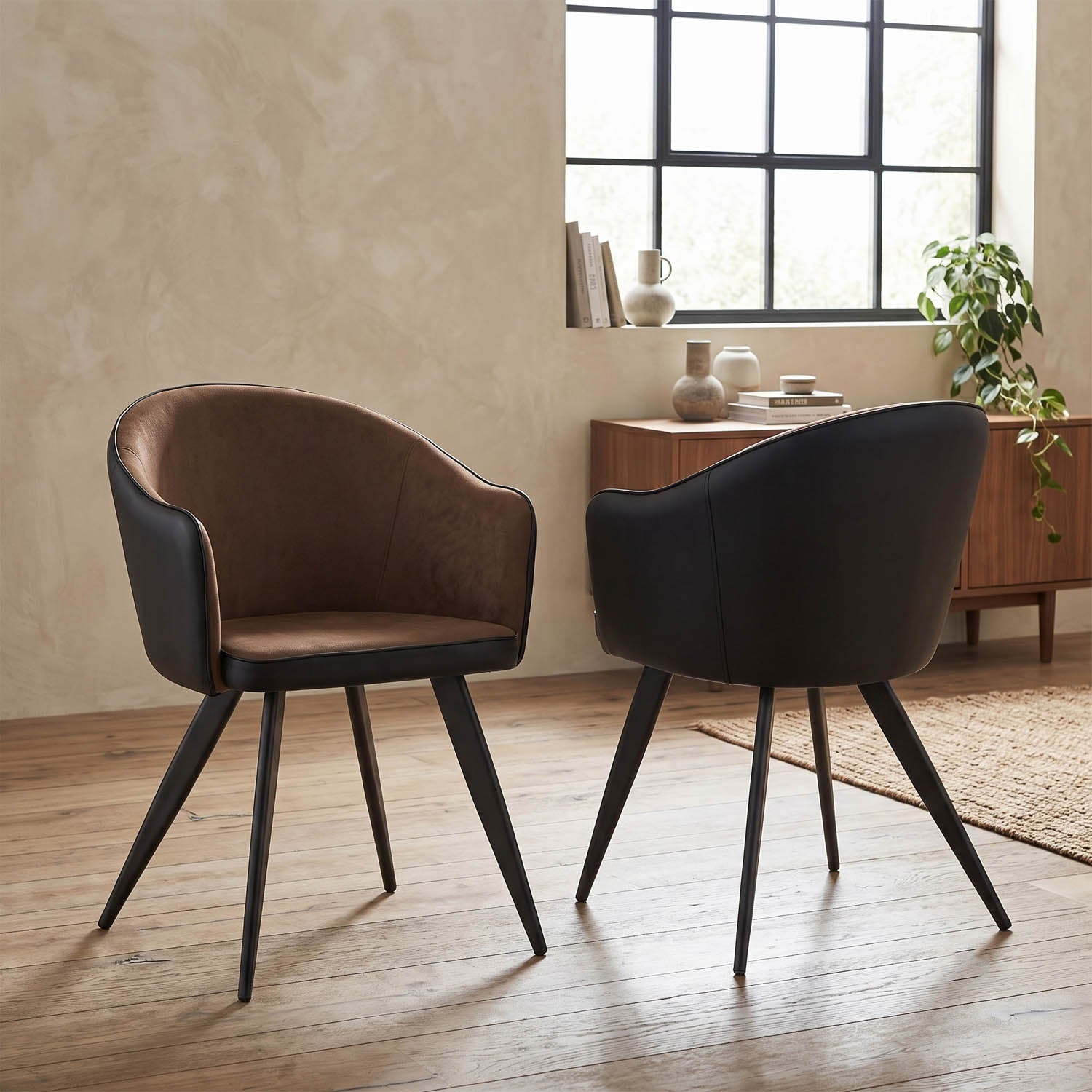 Two modern bucket-style dining chairs with dark brown and black upholstery, shown in a styled room with a wood floor, textured wall, window, and sideboard.