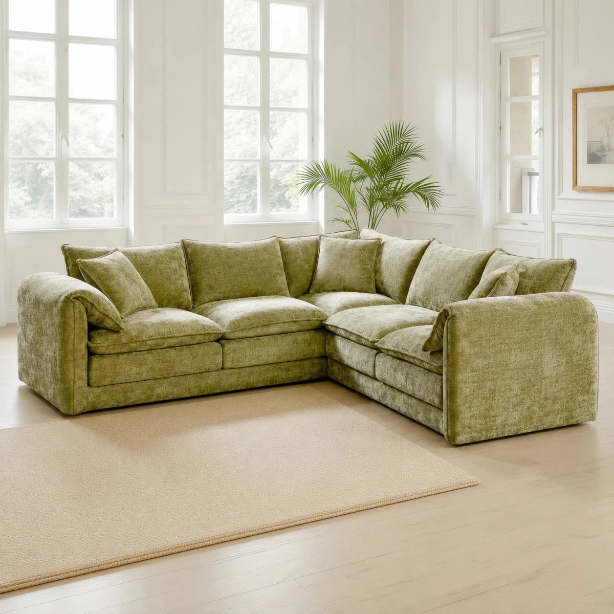 An L-shaped sectional sofa upholstered in textured olive green fabric, styled in a bright, modern living room with white walls, large windows, a light wood floor, and a beige rug.