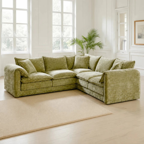 An L-shaped sectional sofa upholstered in textured olive green fabric, styled in a bright, modern living room with white walls, large windows, a light wood floor, and a beige rug.