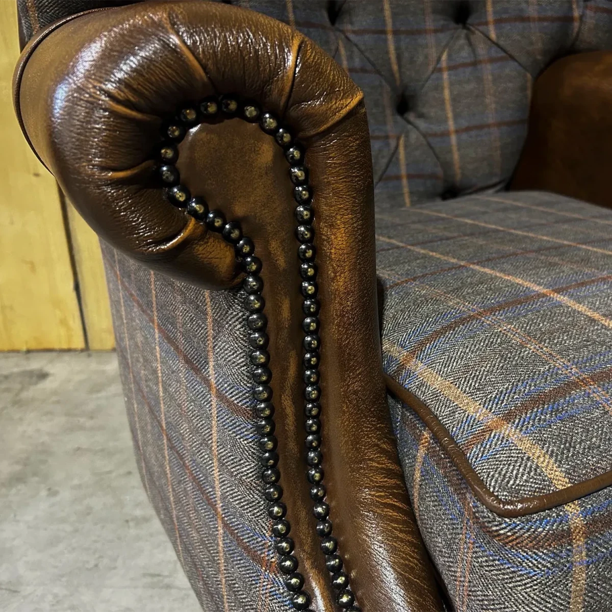 Close-up of a brown leather armrest with decorative nailhead trim on a plaid upholstered chair.
