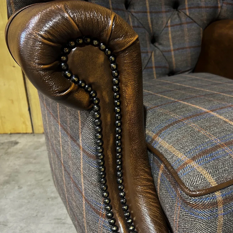 Close-up of a brown leather armrest with decorative nailhead trim on a plaid upholstered chair.