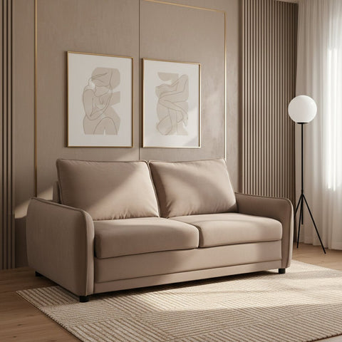 A two-seater sofa bed upholstered in a taupe-beige microfiber fabric, styled in a bright, modern living room with a light rug, floor lamp, and white bookshelf.