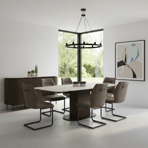 A modern dining room setup featuring a dark brown ribbed rectangular table with a light stone top, surrounded by six taupe upholstered chairs with black metal sled bases. The room has white walls, a large black-framed window looking out to green trees, a black multi-light chandelier, and a matching dark brown ribbed sideboard with small potted plants.