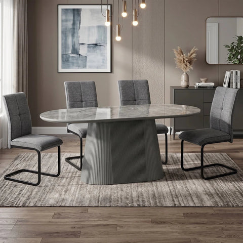 An oval dining table with a ribbed pedestal base and a light gray stone-look top, surrounded by four gray upholstered chairs with black metal legs, set on a textured rug in a modern dining room.
