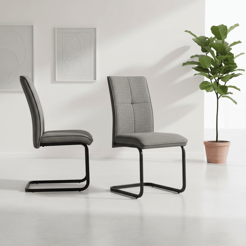 Two modern gray upholstered dining chairs with black cantilever black metal bases, shown in a bright minimalist room with a potted plant and framed wall art.